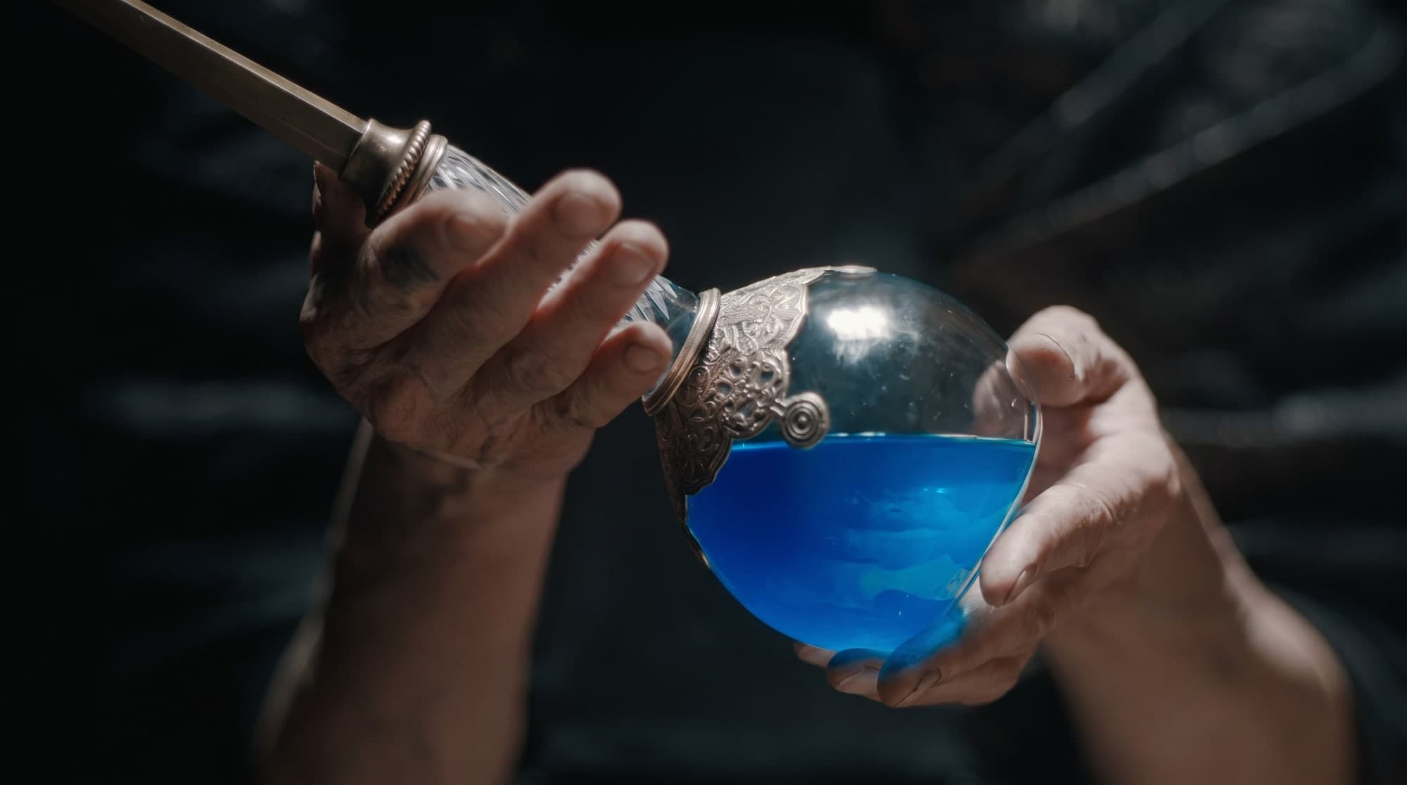 Hands cradling a glass orb of glowing blue elixir in darkness, the Water of Life